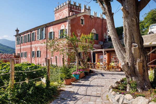 A red building with a balcony, potted plants, and a tree in the foreground.