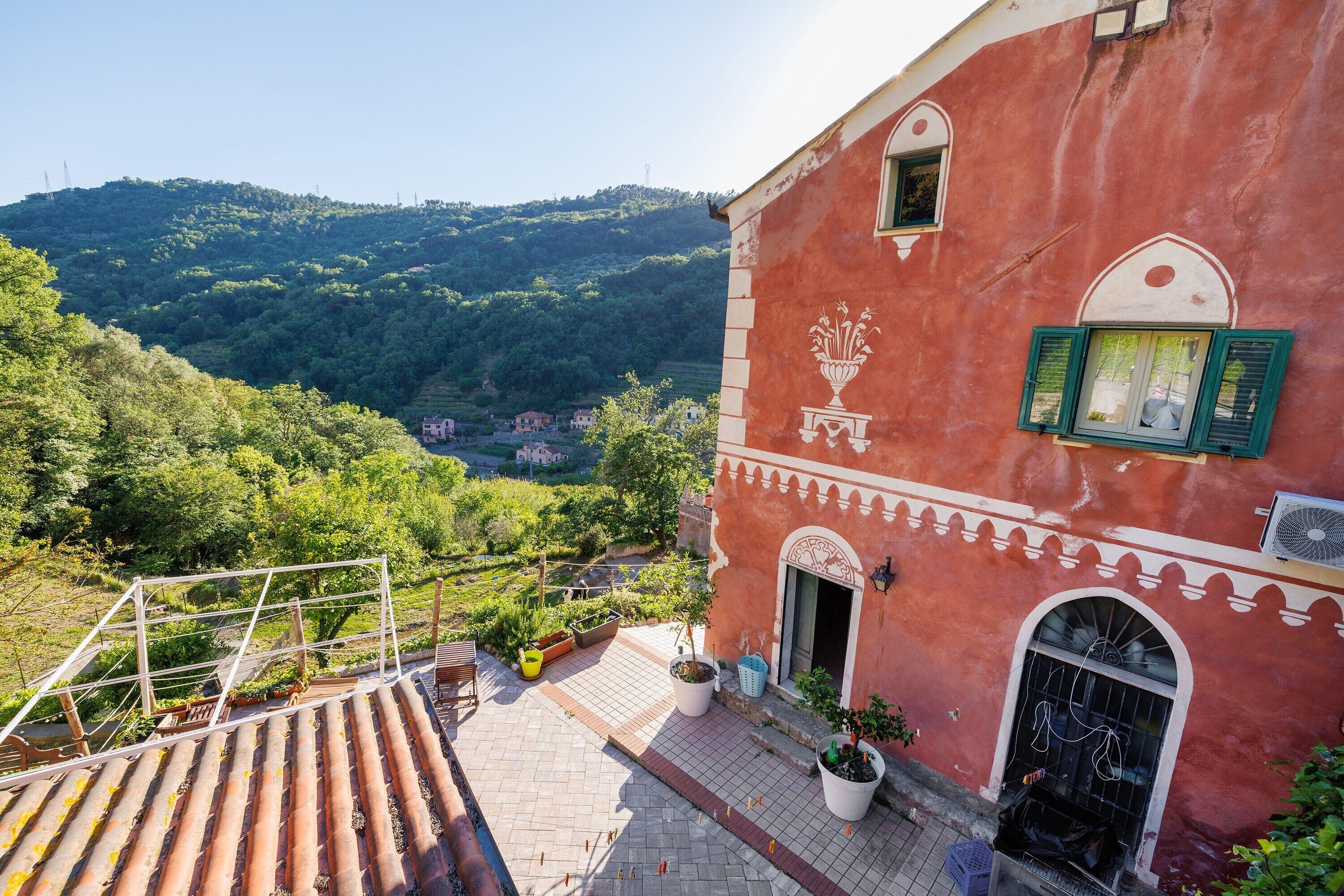A red building with a balcony, potted plants, and a view of a green valley.