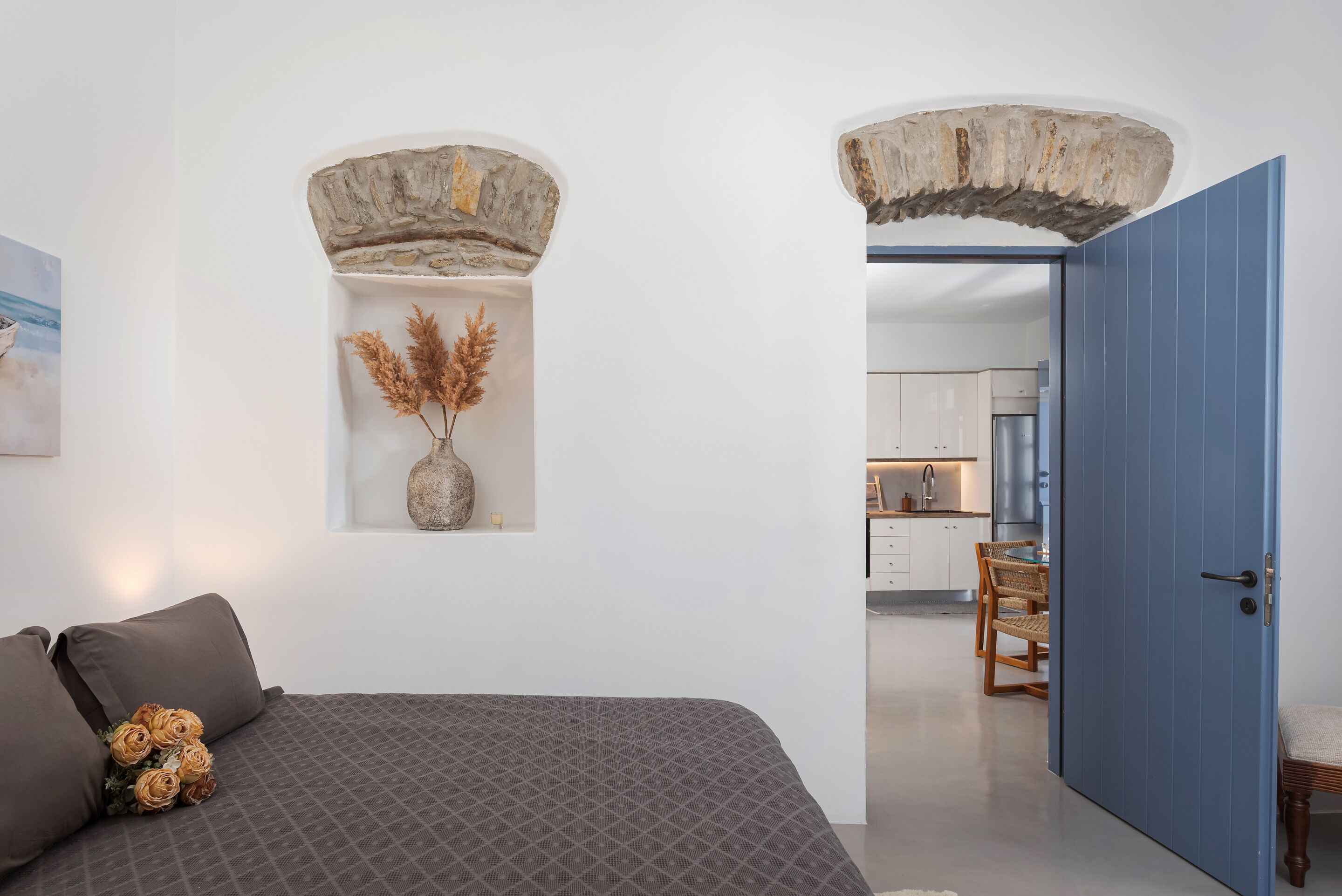 Een slaapkamer met een bed, kussens, een vaas met gedroogde planten en een stenen boog die naar een keuken leidt.