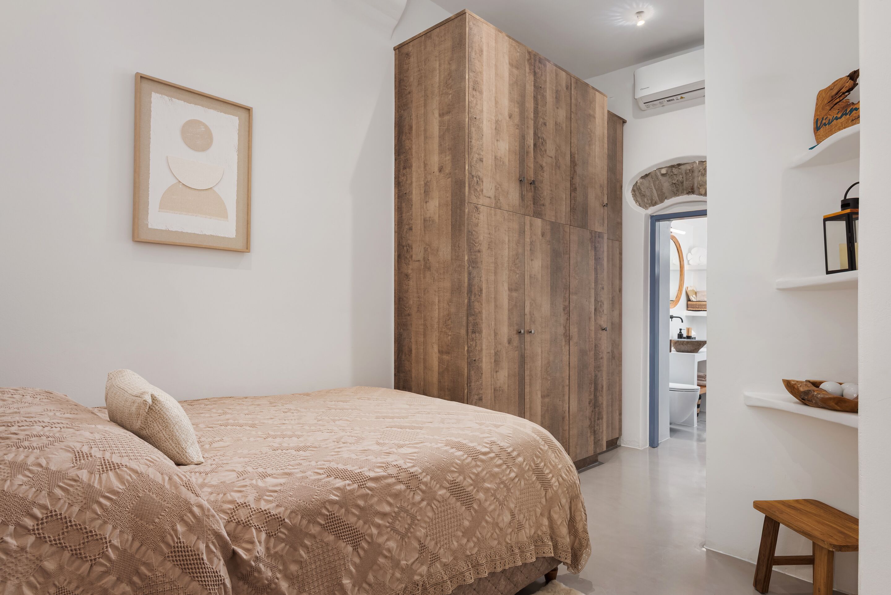 A bedroom with a bed, a wooden wardrobe, a framed picture on the wall, and a bathroom visible through an archway.