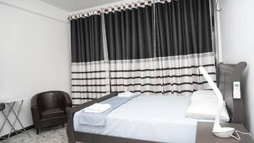 A hotel room with a bed, a chair, a bedside table with a lamp, and a window with curtains.