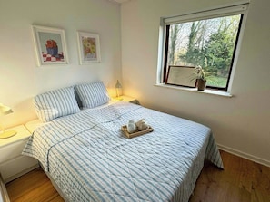 Een slaapkamer met een bed, bedlampen, een raam met een plant en twee foto’s in een lijst aan de muur.
