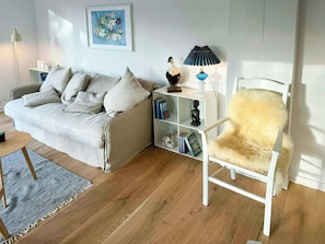 Een woonkamer met een bank, een witte fauteuil, een boekenkast, een lamp en een schilderij aan de muur.