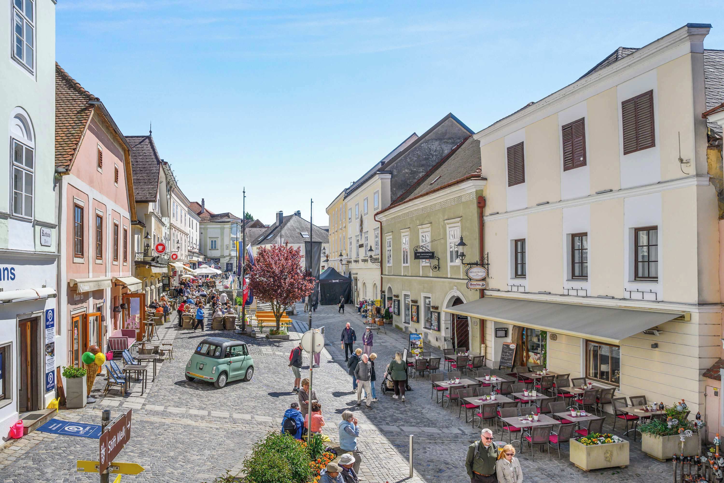 A cobblestone street with outdoor seating, buildings with signage, and a vintage car.