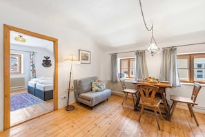 Une pièce comprenant un canapé, une table à manger avec des chaises, un luminaire suspendu et une fenêtre avec des rideaux.
