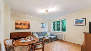 A room with a dining table, chairs, a sofa, a small side table, and a window with a view of trees.