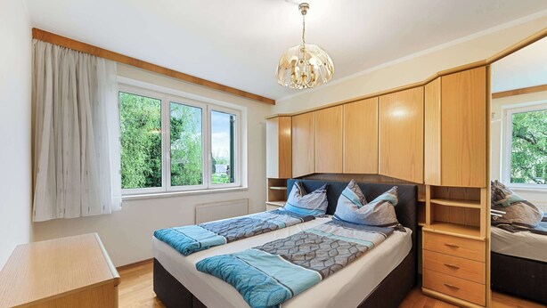 A bedroom with two beds, a wooden wardrobe, a ceiling light, and a window with a view of greenery.