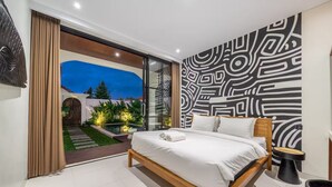 A modern bedroom with a large bed, a bathroom with a black sink, and a view of a garden and pool at dusk. - Alisa 4 Villa (Kerobokan)