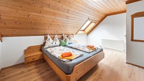 Un dormitorio con techo inclinado, suelo de madera, cama con almohadas y manta, mesita de noche y tragaluz.