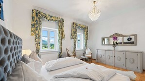 Ein Schlafzimmer mit einem großen Bett, zwei Fenstern mit floralen Vorhängen, einer Kristallleuchtre, einem Schminktisch und einem Spiegel.