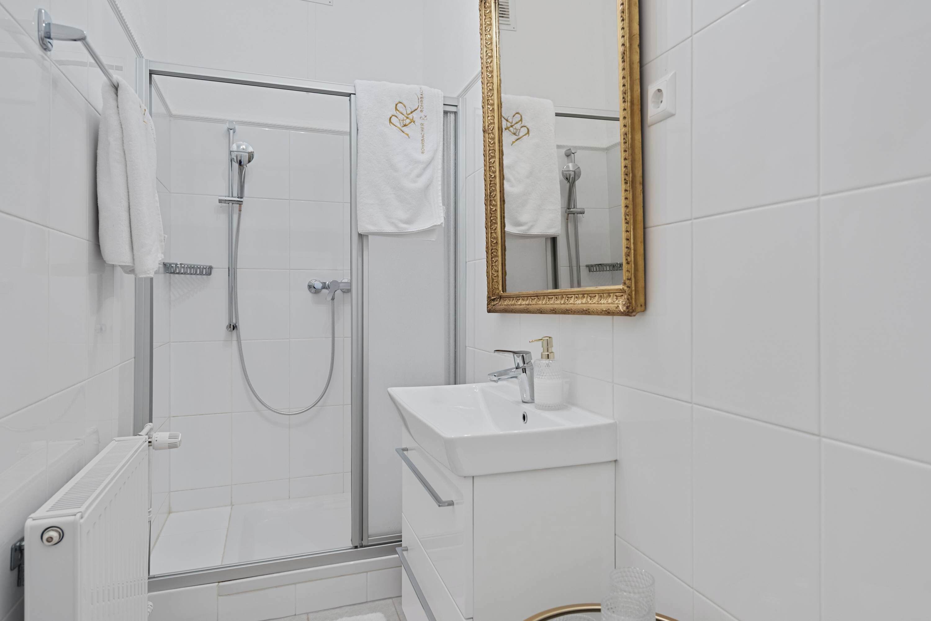 Et moderne badeværelse med en glasbrusekabine, en hvid vask og et spejl med gylden ramme.