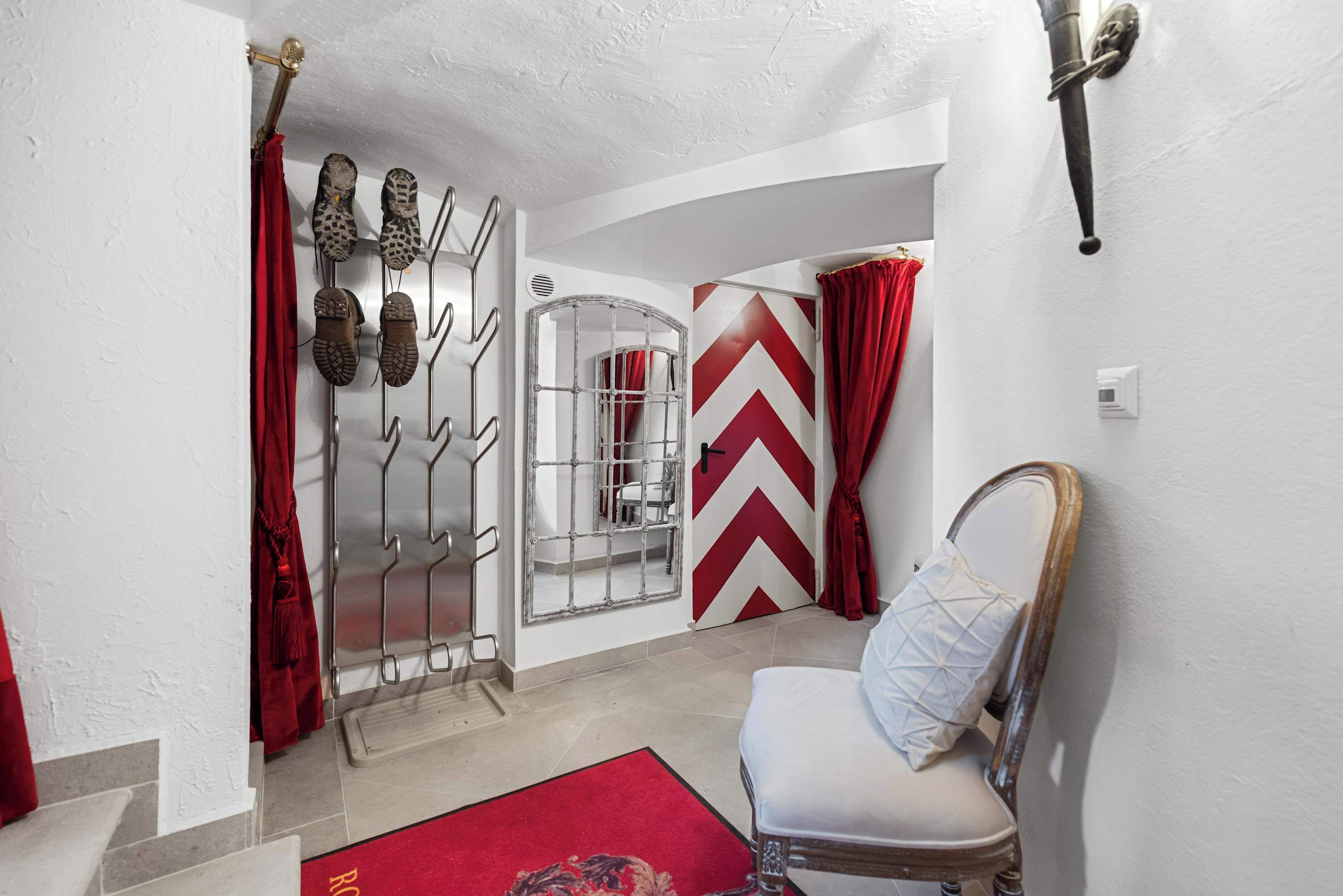 Quarto com uma cama, uma cadeira, parede com padrão geométrico vermelho e branco, cortina vermelha e sapateira com sapatos.