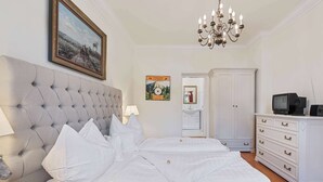 A bedroom with a tufted headboard, a bed with white linens, a chandelier, a television, and a wardrobe.