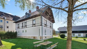 A two-story building with a sloped roof, a stone foundation, and a small garden with a bench and a table.