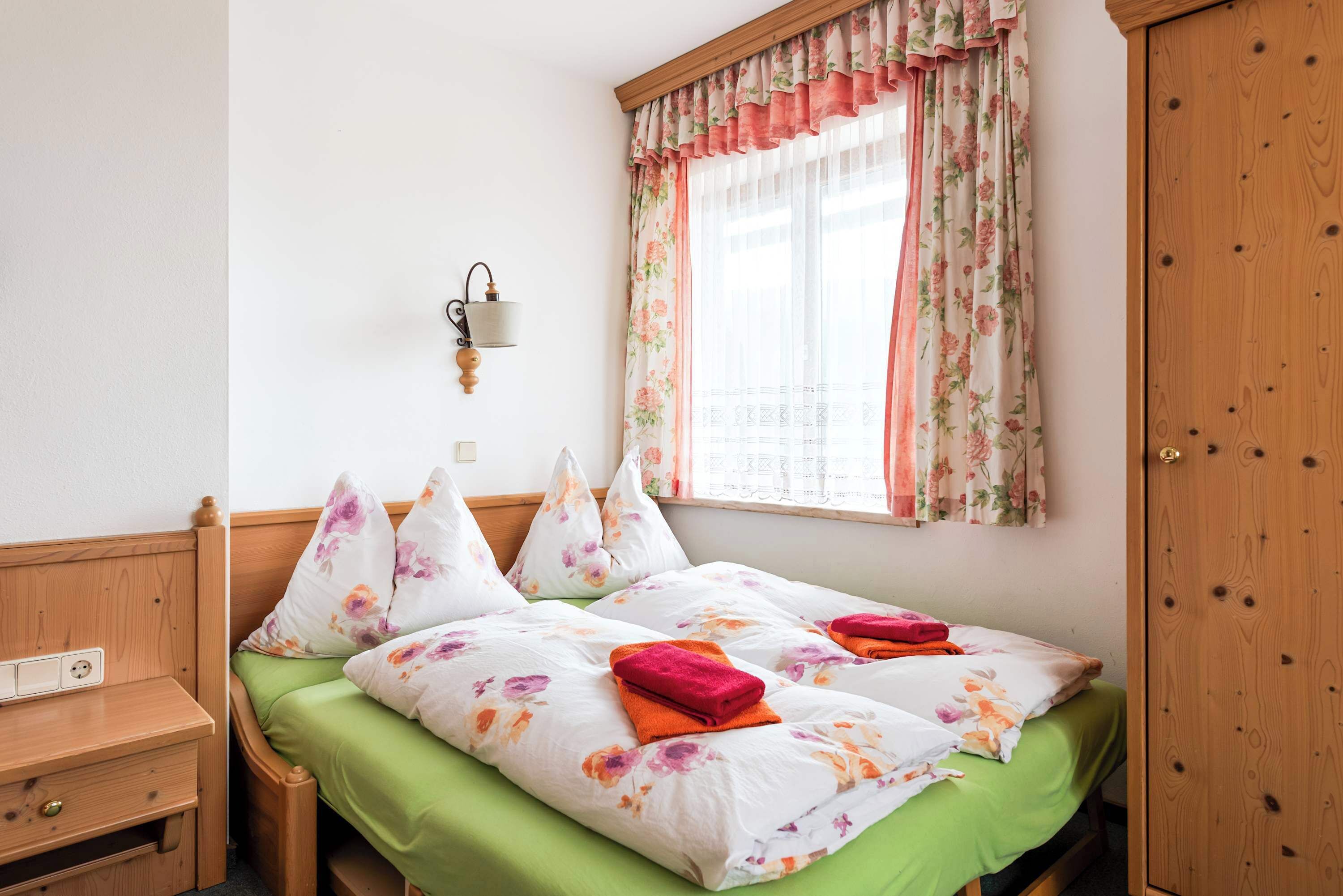 Une chambre avec une tête de lit en bois, une table de chevet avec des tiroirs, une couvre-lit verte, des coussins à motifs floraux et une fenêtre ornée de rideaux à fleurs.