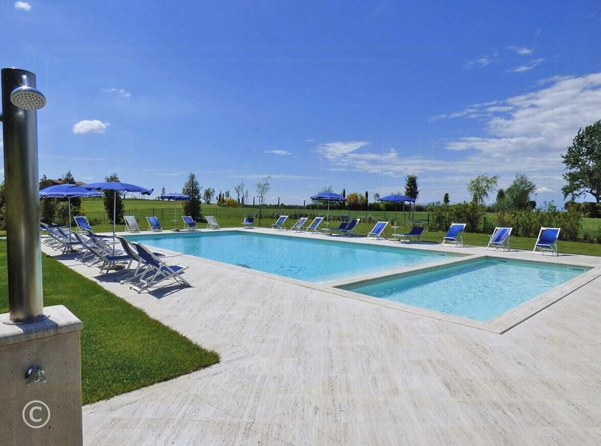 Una piscina con diversi lettini e ombrelloni, immersa nel verde e circondata da alberi.