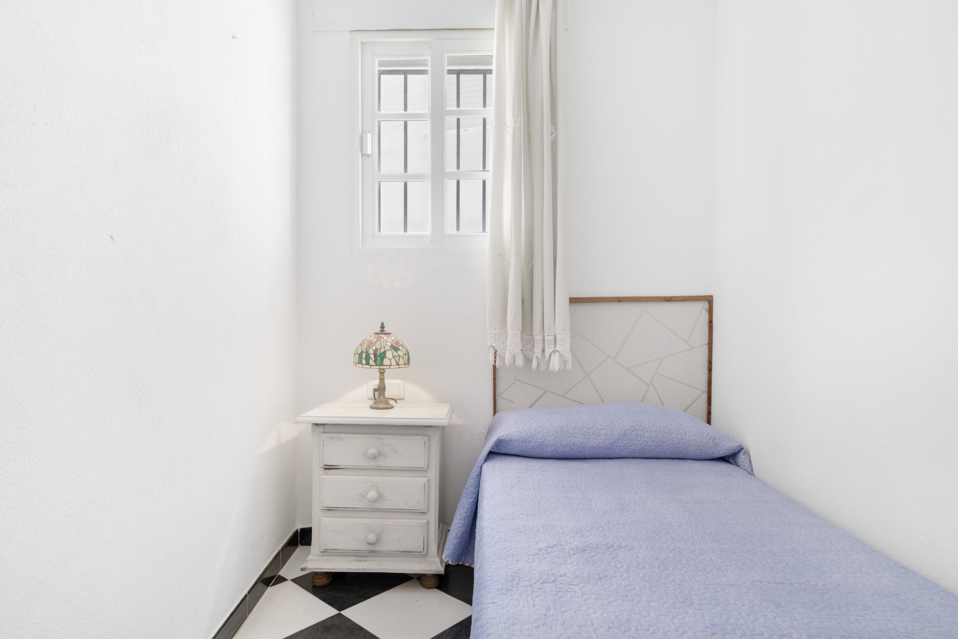 A small, white room with a bed, a nightstand with drawers, a window with white curtains, and a geometric-patterned headboard.