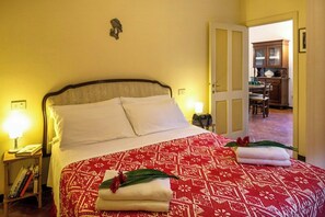 Un lit bien fait, recouvert d’une couvre-lit à motifs, une lampe de chevet et une petite table avec des livres et une autre lampe.