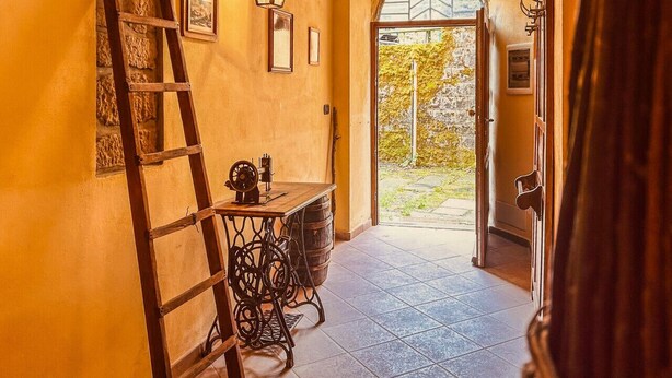 Un couloir avec une échelle en bois, une table surmontée d’un tonneau et une porte donnant sur l’extérieur.
