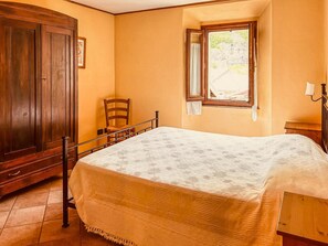 A bedroom with a bed, a wooden wardrobe, a chair, a window with curtains, and a nightstand.