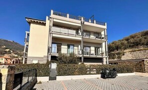 A multi-story building with balconies, a paved entrance area, and a surrounding landscape. - Hostdomus - Vittorio House (Finale Ligure)