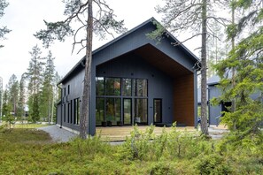 A modern two-story building with large windows, a wooden deck, and a surrounding forest.