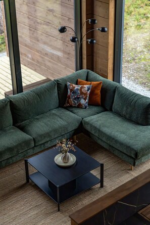A green sectional sofa with a decorative pillow, a small round table with a vase, and a modern light fixture.