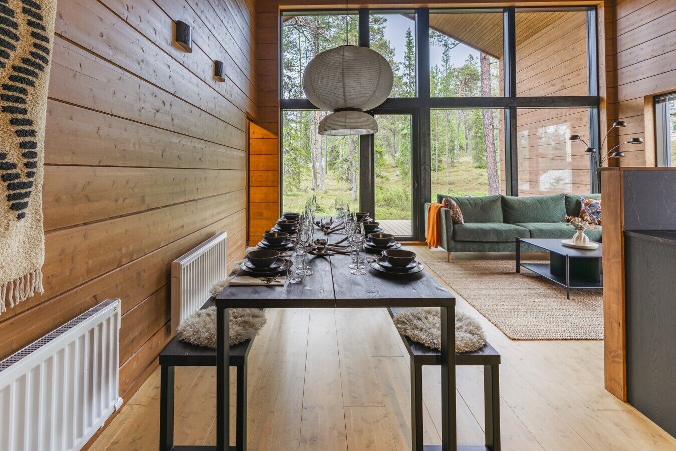A dining area with a long table set for a meal, a sofa, and a view of the forest through large windows.