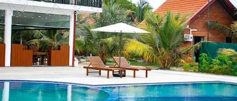 A swimming pool with lounge chairs and umbrellas, surrounded by palm trees and a building with a red roof.