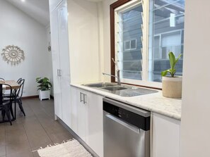 A modern kitchen with white cabinets, a stainless steel dishwasher, a window with frosted glass, and a potted plant. - Mahalo at Anna Bay (Anna Bay)