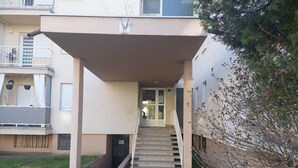 Ein Gebäudeeingang mit einer überdachten Treppe, die zu einer Tür führt, eine kleine Grünfläche mit Gras und ein Baum zur Rechten.