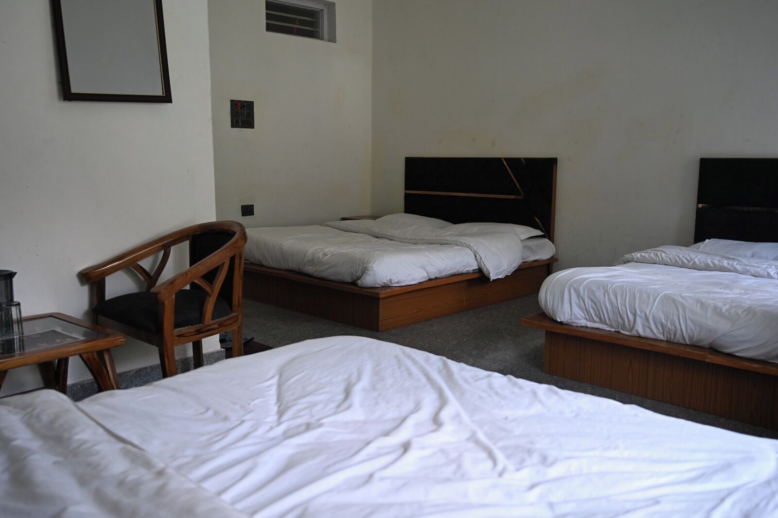 Quarto com três camas, uma cadeira de madeira, uma mesa pequena e um painel de controle fixado na parede.