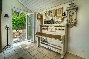 Una habitación con una puerta de vidrio que da al exterior, un estante blanco con relojes y botellas, y una pequeña mesa con botellas.