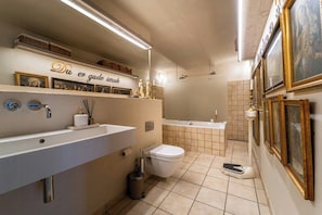 Ein Badezimmer mit einem großen Waschbecken, einer Toilette und einer Badewanne. An den Wänden hängen Bilder in Rahmen und ein Regal mit Dekorationsobjekten.