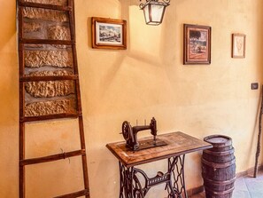 Una máquina de coser antigua sobre una mesa de madera, una escalera de madera y un barril frente a una pared texturizada con cuadros enmarcados.