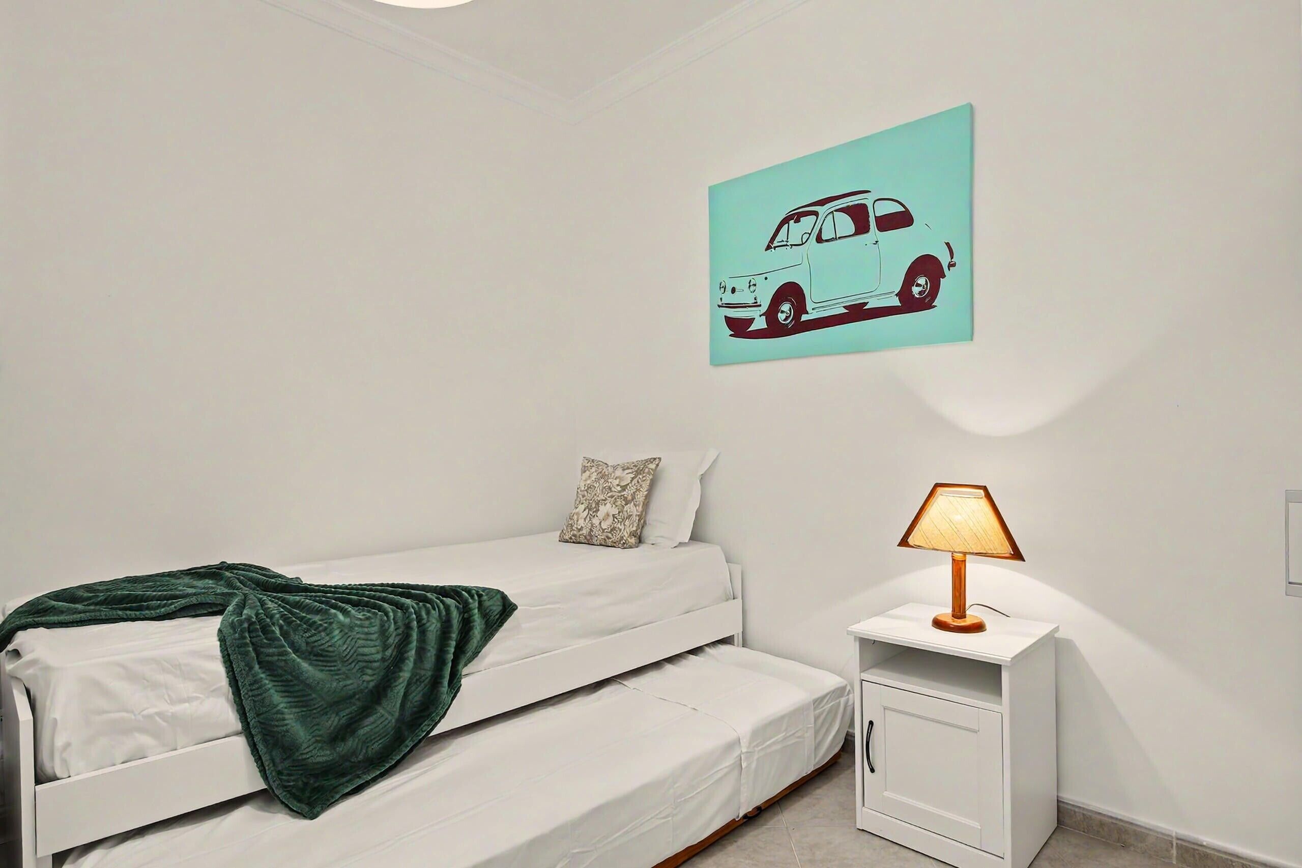 A bedroom with a bunk bed, a bedside table with a lamp, a wall-mounted car decal, and a small cabinet.