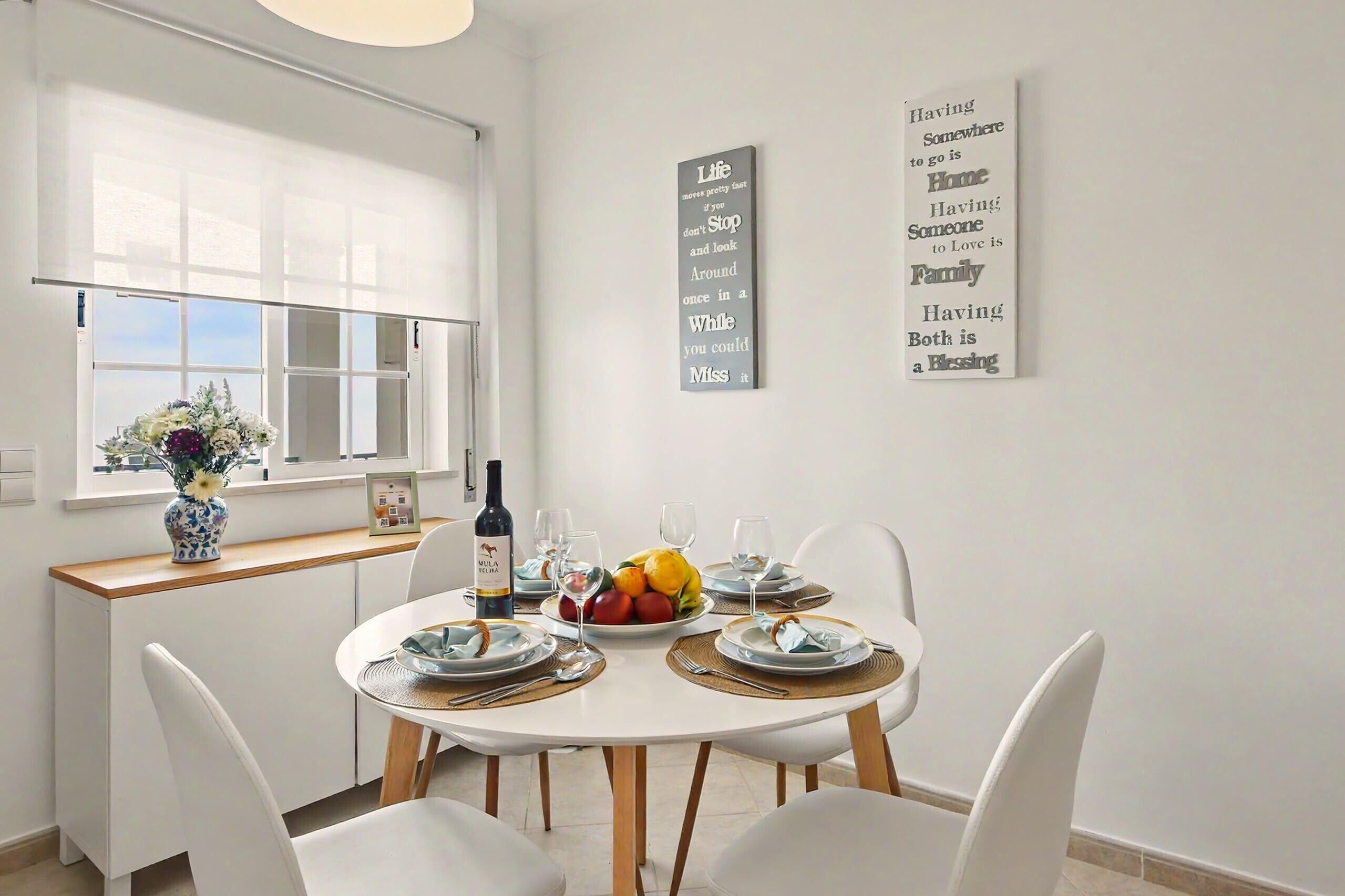 Comedor con una mesa redonda para cuatro personas, sillas blancas, un jarrón con flores y un frutero.