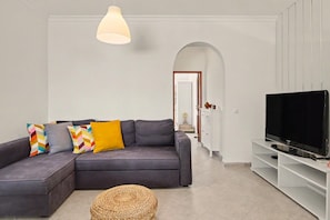 A living room with a grey sofa, colorful cushions, a woven ottoman, a TV on a white stand, and a pendant light. - Sun & Green Apartment – Near Tavira and the Beach (Conceição)