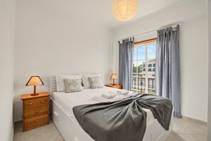 Ein Schlafzimmer mit einem Bett, zwei Nachttischen, einer Lampe, einem Fenster mit Vorhängen und Blick auf Gebäude.