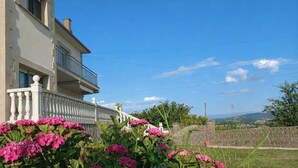 En tvåvåningsbyggnad med en balkong, omgiven av frodig grönska och rosa blommor.
