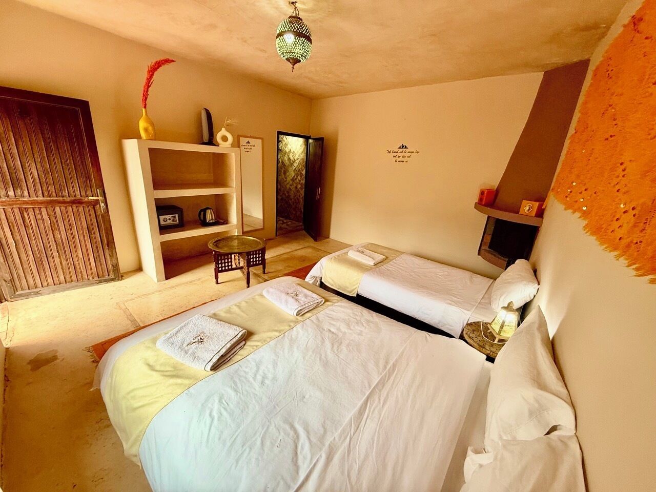 Una habitación con dos camas, una mesita, una estantería con objetos decorativos y una puerta.