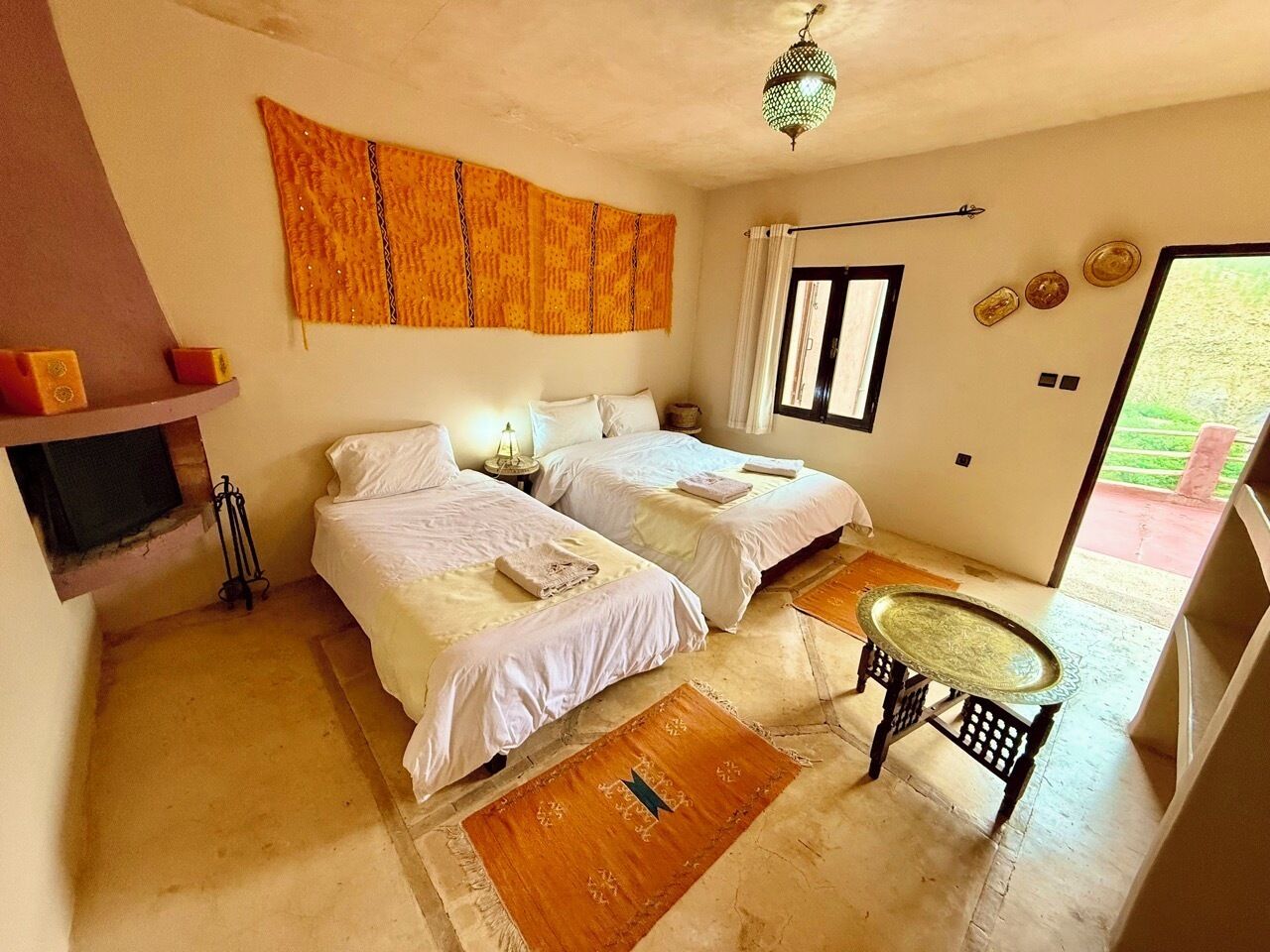 Une chambre avec deux lits, une petite table, une tapisserie murale et une fenêtre avec des rideaux.