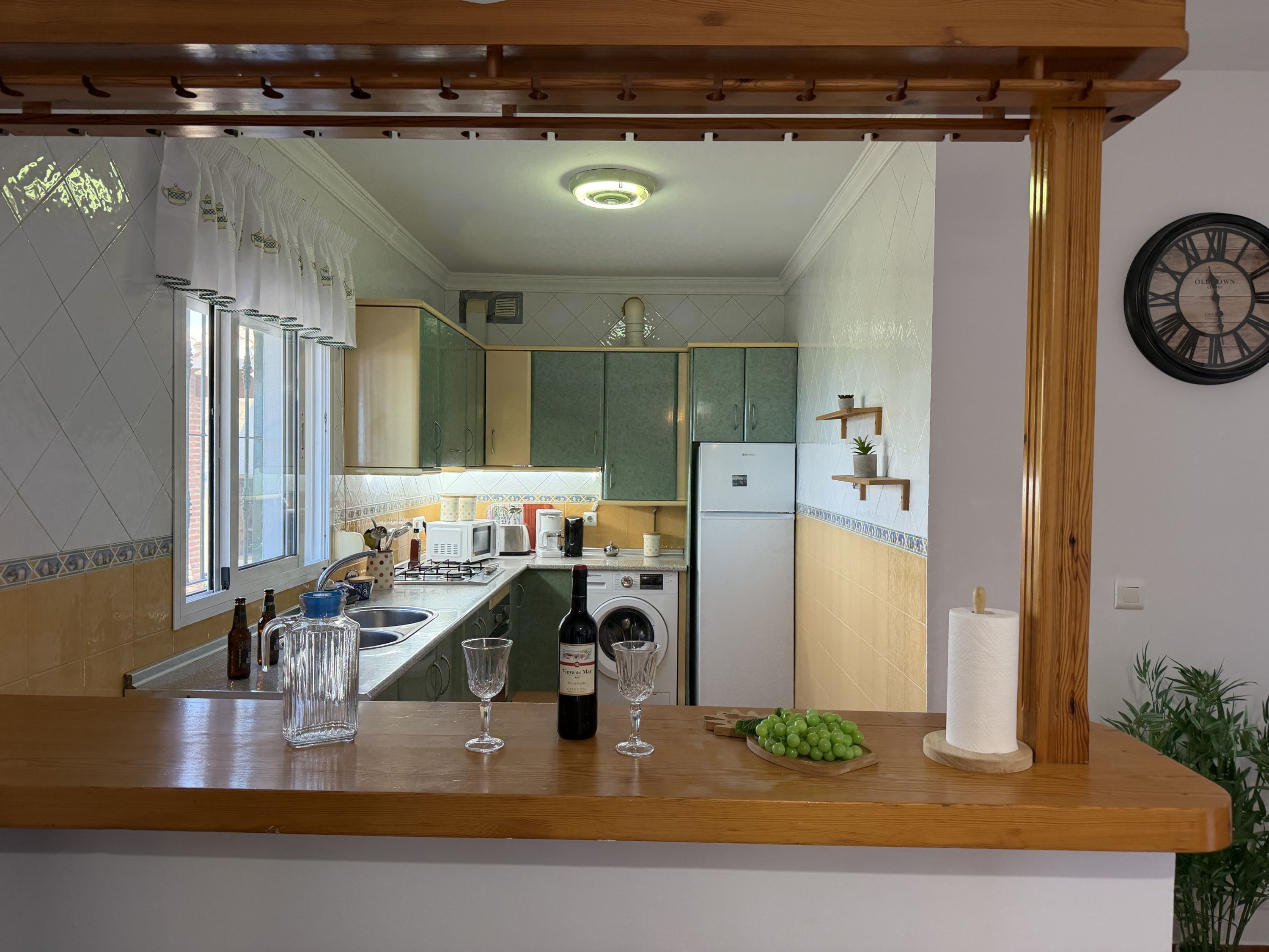 Een keuken met een houten aanrecht, een spoelbak, een wijnfles en een klok aan de muur.