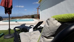 Zona junto a la piscina con sillones de descanso y una sombrilla, una pared blanca y vista al mar.