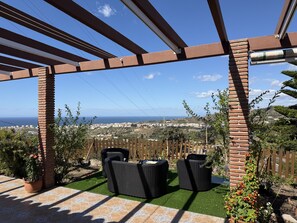 A covered patio with wicker furniture, a brick pillar, and a view of the sea and cityscape. - Cortijo del Verdial by At Home Costa del Sol (Torrox)