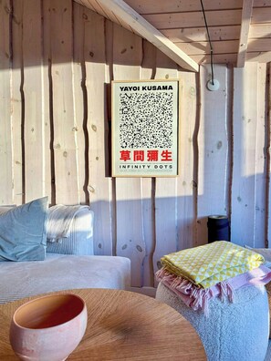 L’intérieur d’une cabane en bois, avec un lit, des oreillers, une table sur laquelle se trouve un bol, et une œuvre d’art encadrée accrochée au mur.