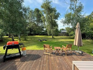 Une terrasse dotée de mobilier d’extérieur, d’un barbecue et offrant une vue sur une cour verdoyante bordée d’arbres.