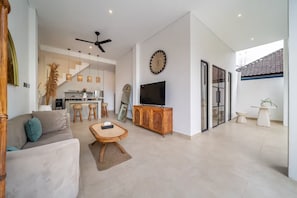 Una sala de estar moderna con sofá, una mesa de centro, televisor sobre un mueble de madera, ventilador de techo y una cocina al fondo.
