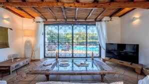 A room with a TV, a table with books, a window with a view of a pool, and a wooden ceiling. - Villa Flor, Exclusive Property With Pool in Stintino (Stintino)