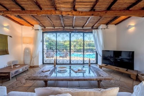 A room with a TV, a table with books, a window with a view of a pool, and a wooden ceiling. - Villa Flor, Exclusive Property With Pool in Stintino (Stintino)